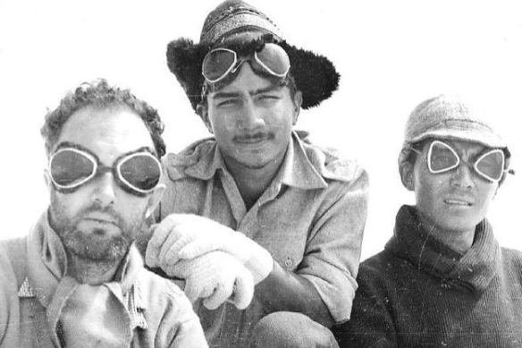 Three individuals in a black and white photo wearing goggles and winter clothing, set against a snowy backdrop.